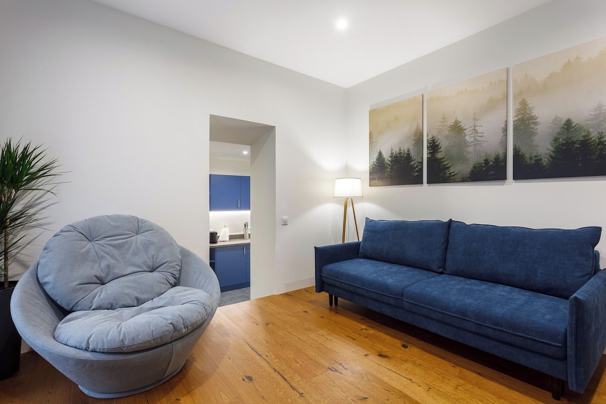 A living room with a grey bean bag chair, a blue sofa, a wooden floor, and a kitchen in the background.