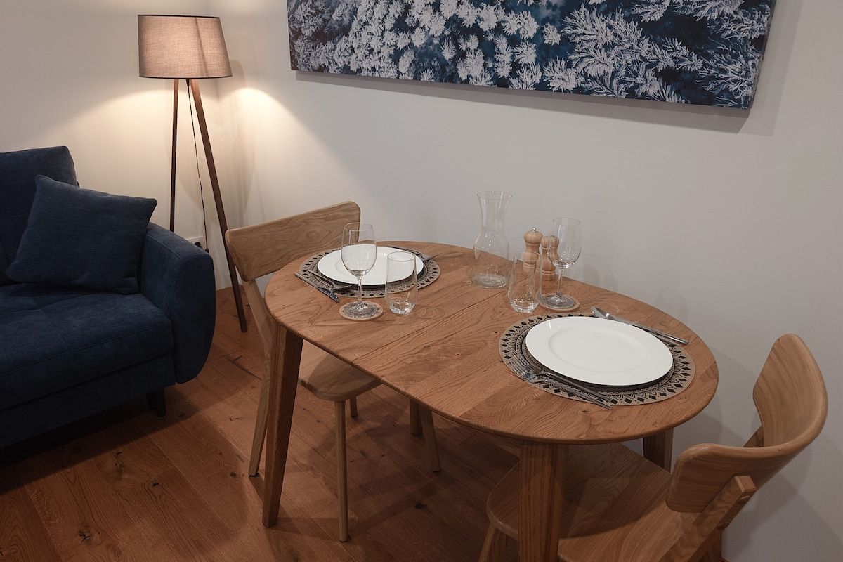 Et spiseområde med et træbord dækket til to personer, to stole, en blå sofa og en gulvlampe.