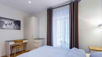 Une chambre d’hôtel comprenant un lit, un bureau avec une chaise, une armoire et une grande fenêtre avec des rideaux.
