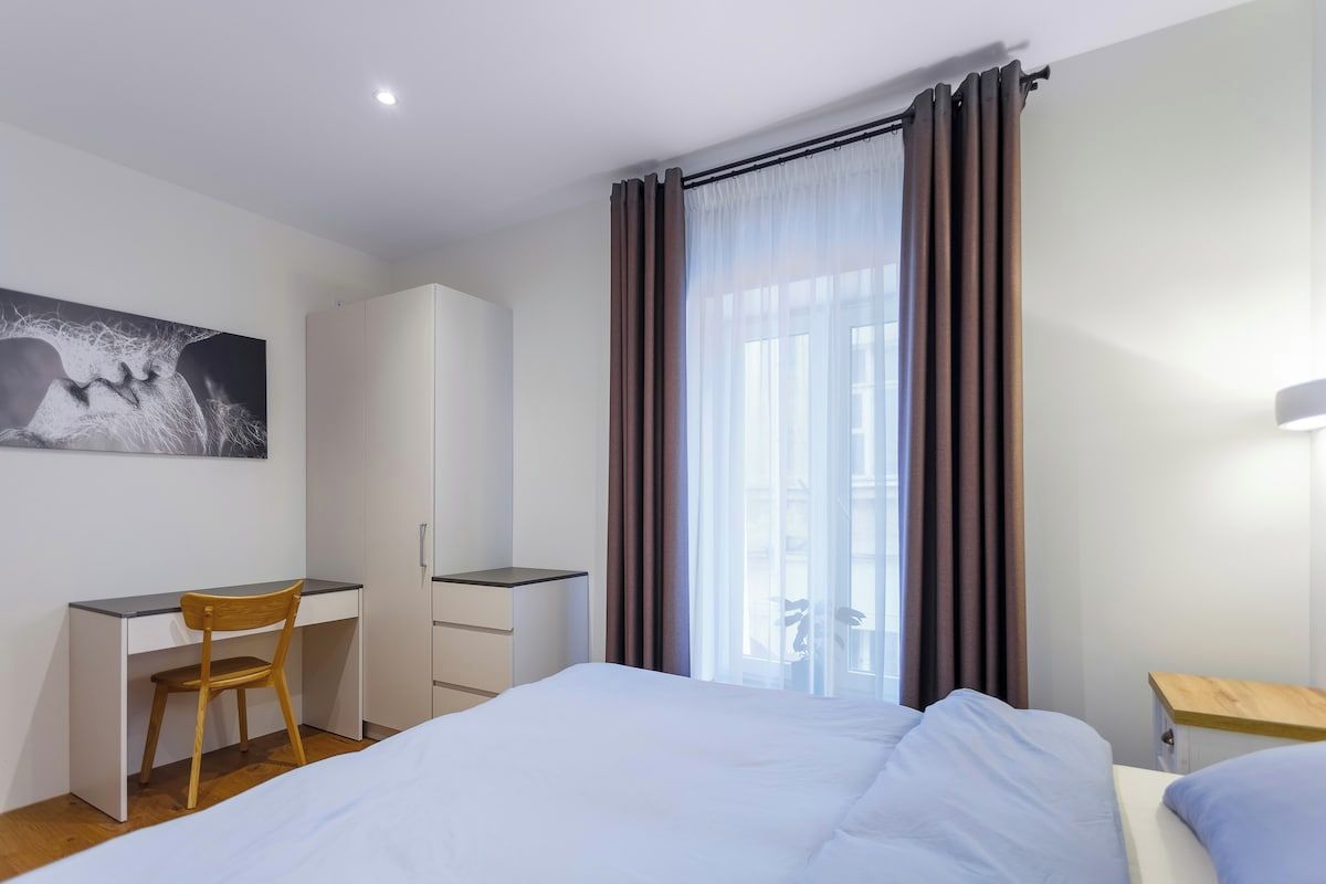 Quarto de hotel com cama, escrivaninha com cadeira, guarda-roupa e uma janela grande com cortinas.