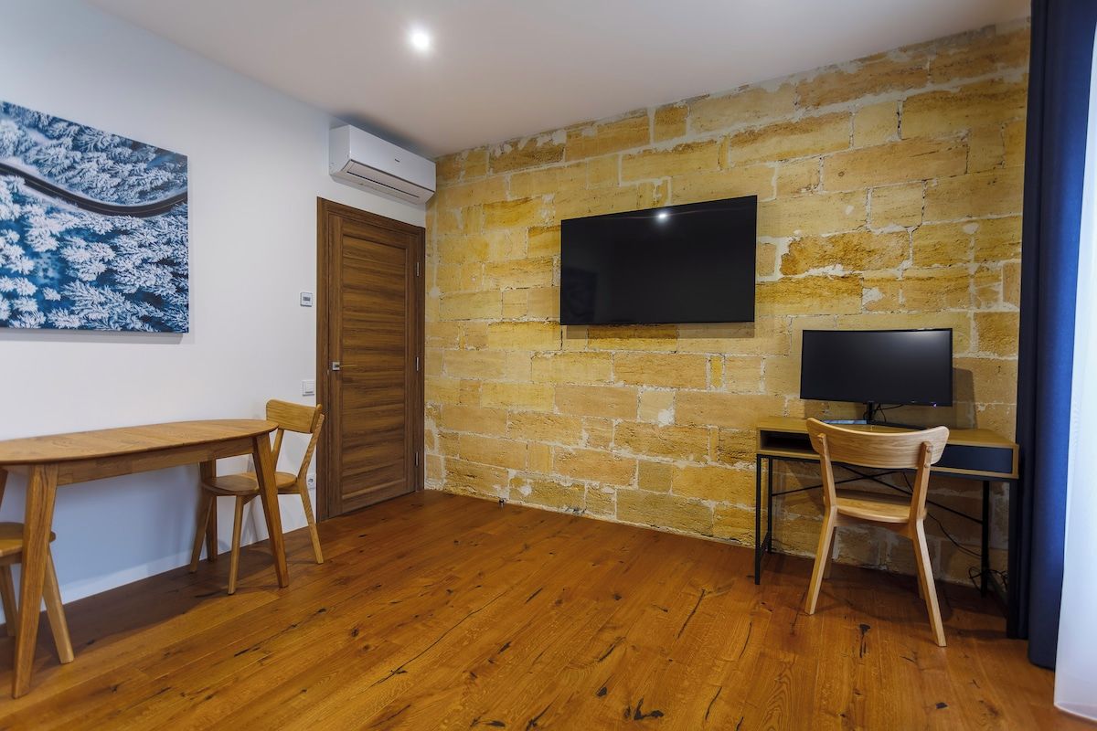 A room with a wooden floor, a stone wall, a flat-screen TV mounted above a desk, and a wooden door.
