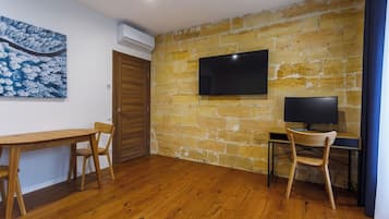 A room with a wooden floor, a stone wall, a flat-screen TV mounted above a desk, and a wooden door.