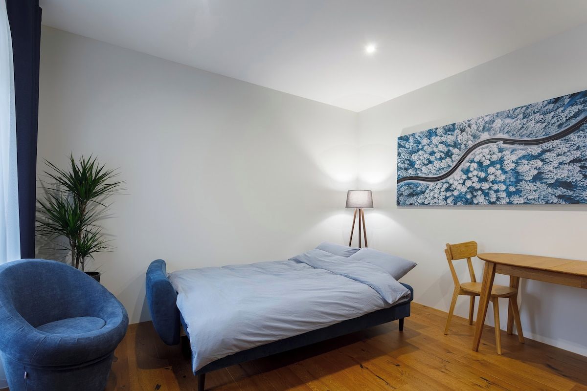 A bedroom with a bed, a desk, a chair, a lamp, and a wall painting.