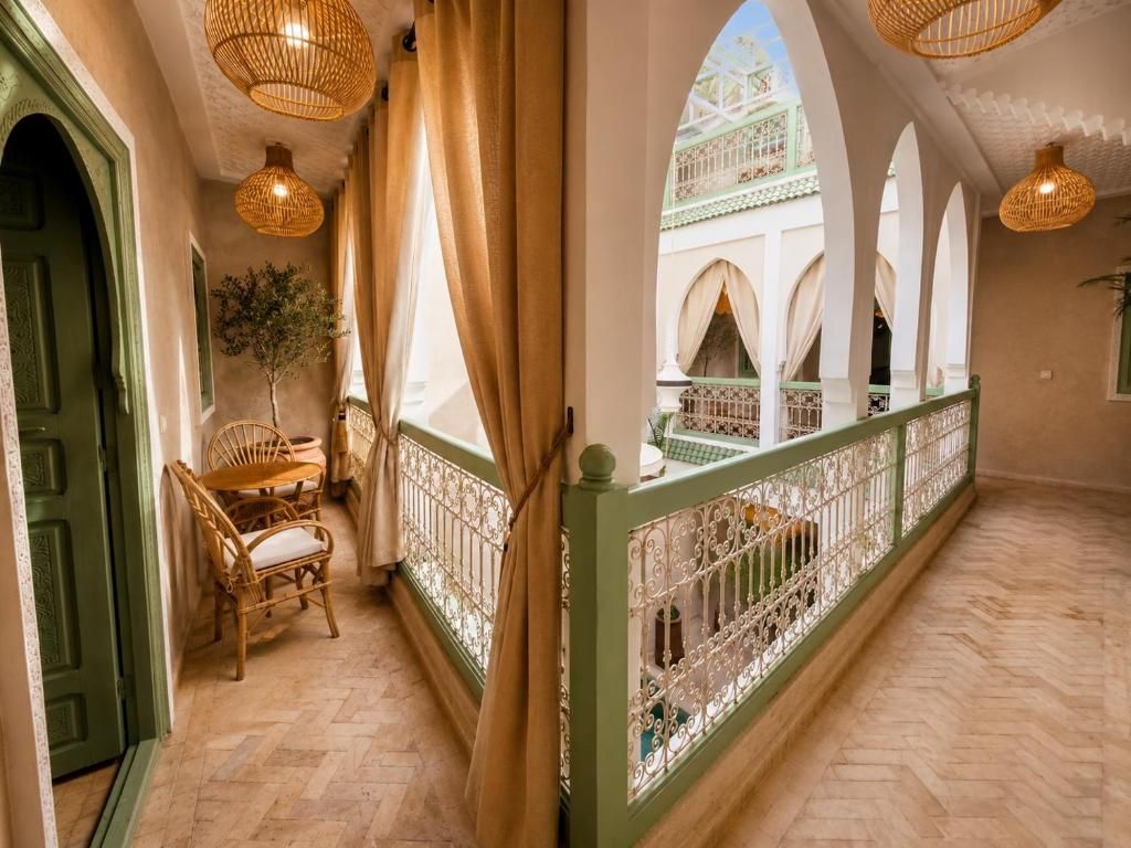 Un couloir avec une porte verte, des chaises en osier, une plante et une vue sur une cour intérieure ornée d’arcades.