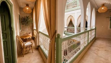 Un couloir avec une porte verte, des chaises en osier, une plante et une vue sur une cour intérieure ornée d’arcades.