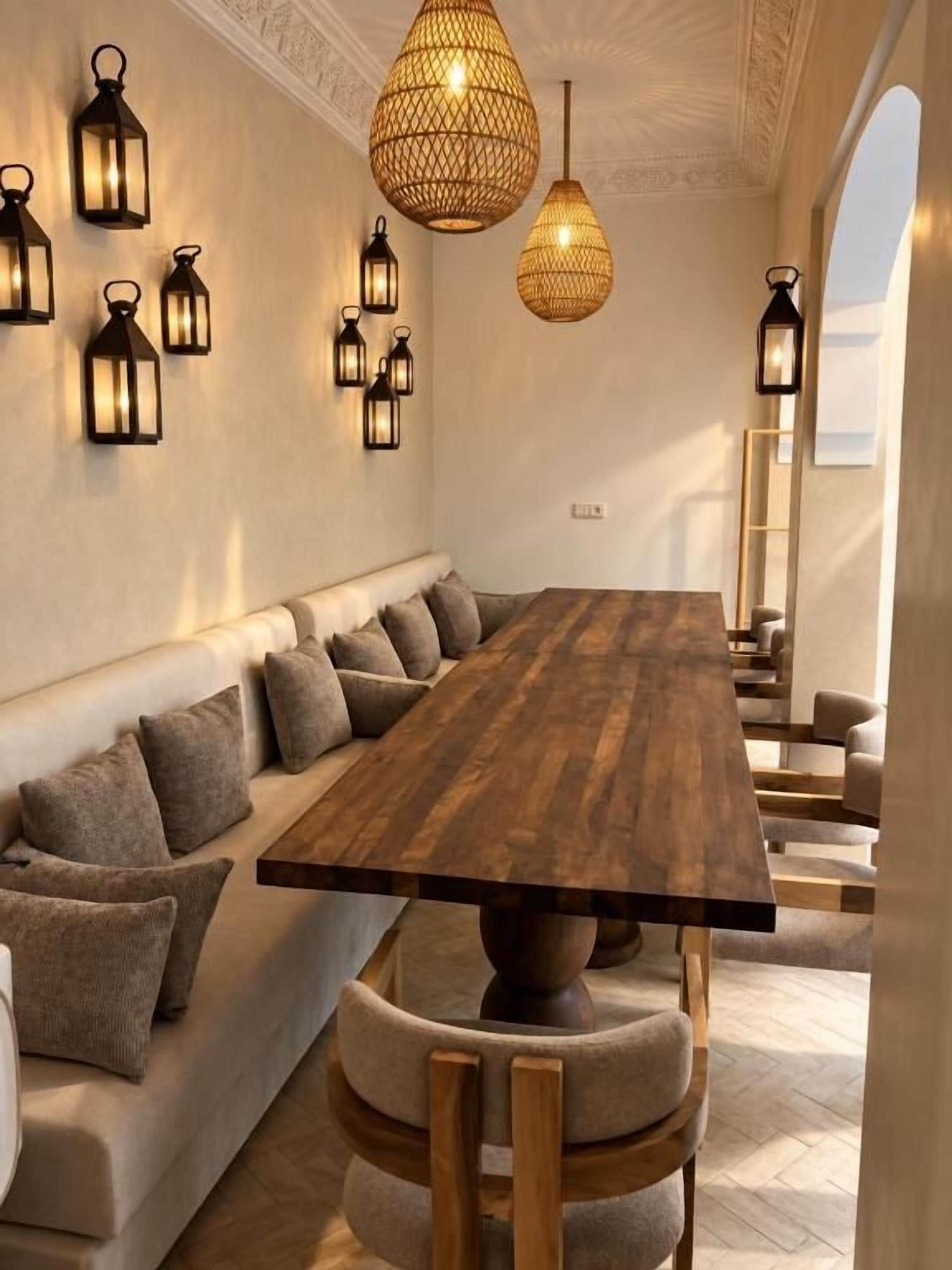 Un espace repas avec une longue table en bois, des sièges rembourrés et des lanternes fixées au mur.