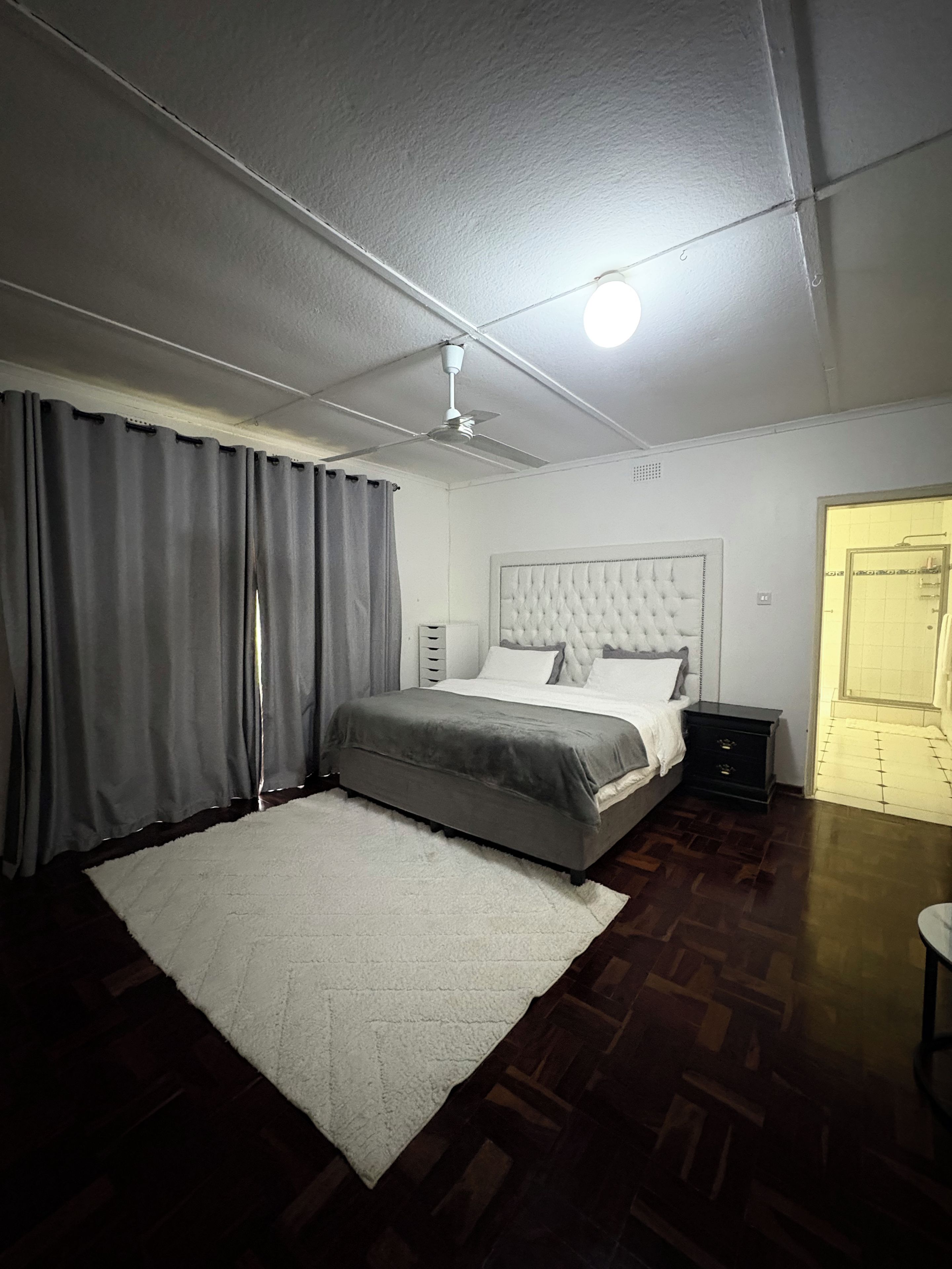 A bedroom with a bed, a bedside table, a nightstand, a ceiling fan, and a door leading to another room.