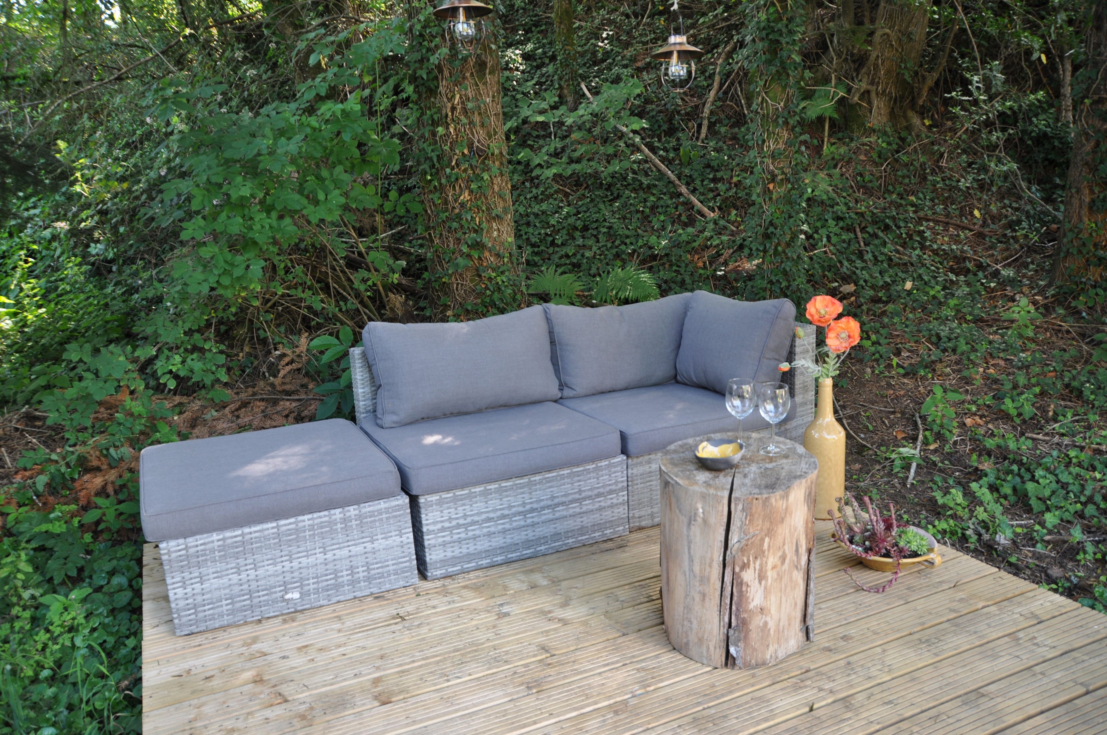 Un canapé d’angle gris est installé sur une terrasse en bois, entouré de verdure.