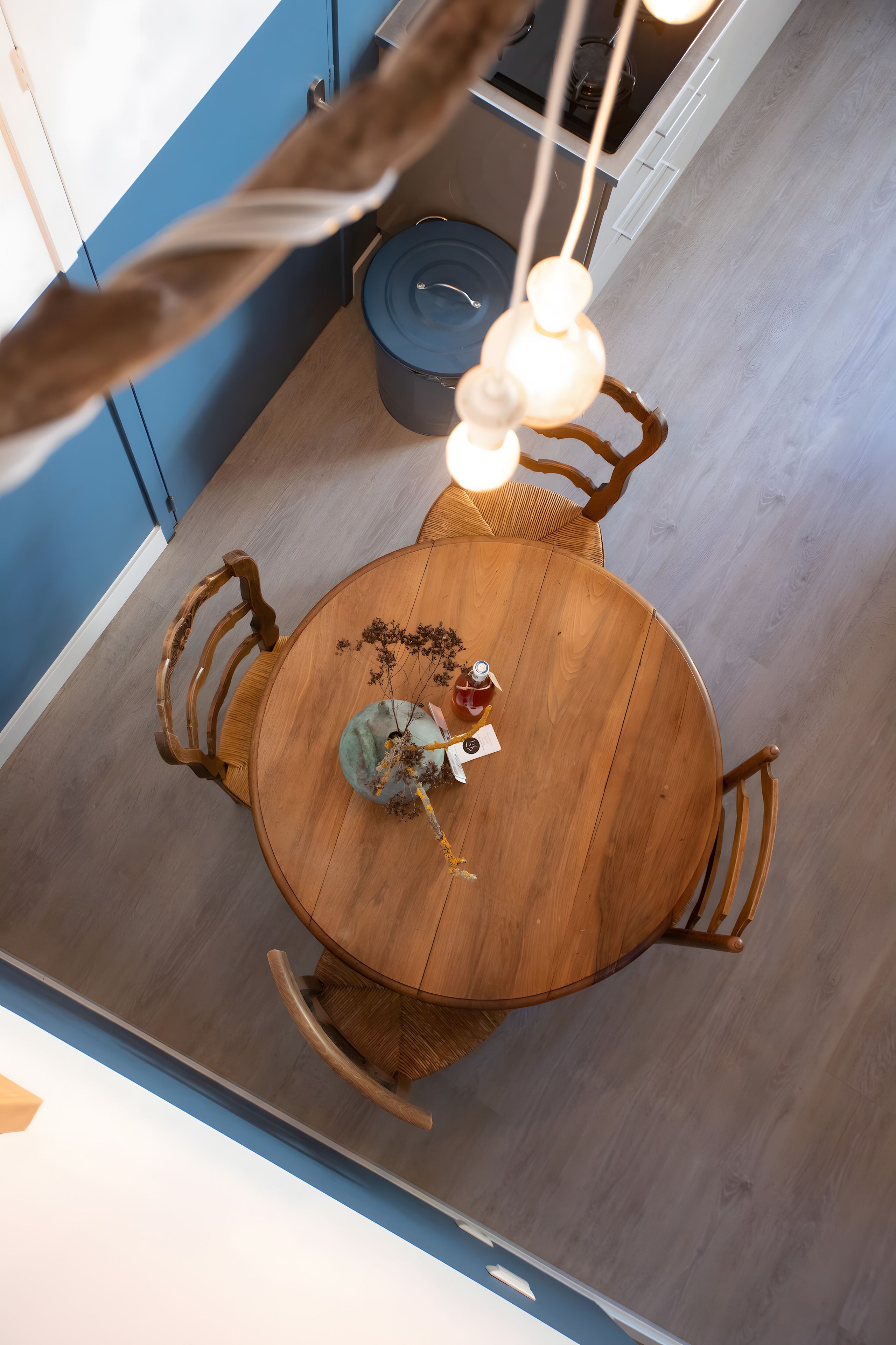 Une table de salle à manger ronde en bois, avec des chaises, un vase contenant des fleurs séchées et une bouteille posée sur la table.
