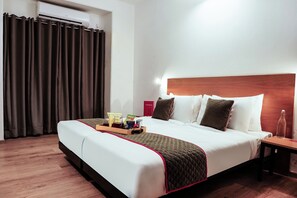 Quarto de hotel com cama, mesa de cabeceira, bandeja com petiscos, uma garrafa e um letreiro vermelho.