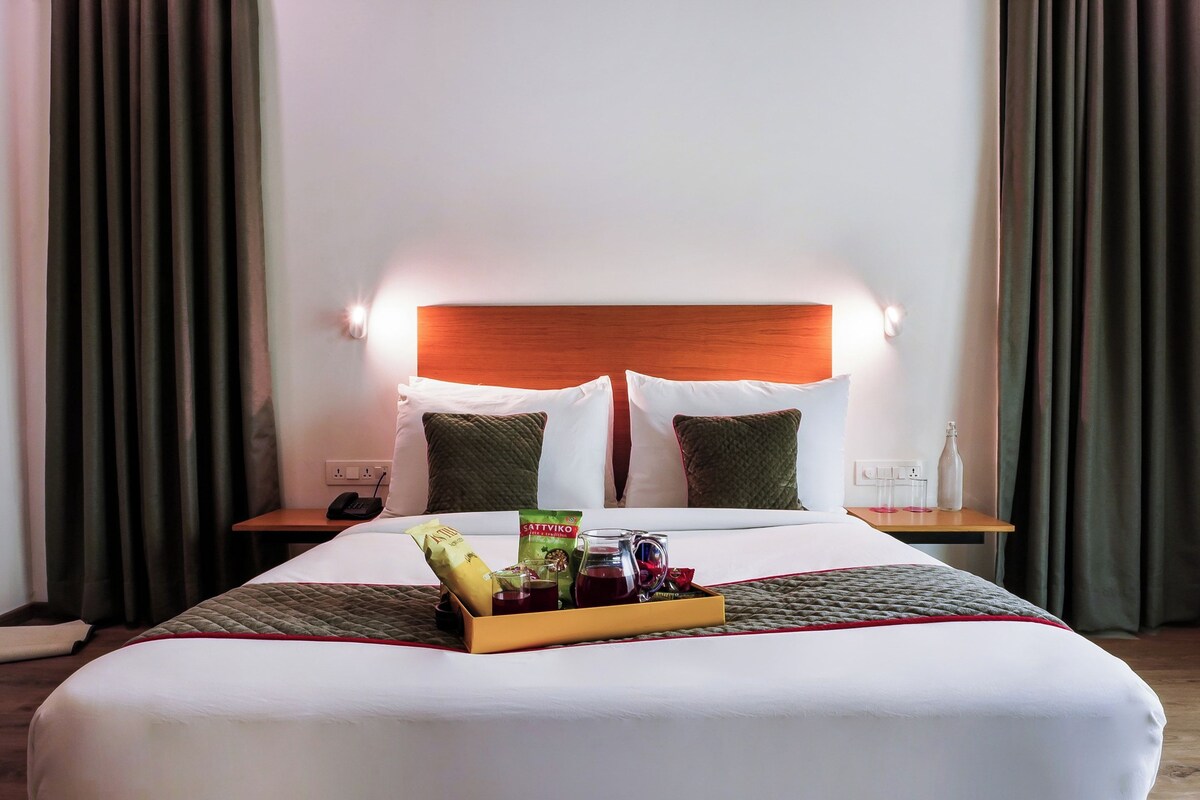 Quarto de hotel com cama, mesa de cabeceira com telefone, bandeja com lanches e uma garrafa de água.