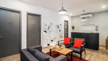 A modern hotel lobby with a reception desk, two seating areas with red cushions, a wall mural, and a wooden coffee table with snacks.
