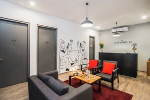 Um hall de hotel moderno com balcão de recepção, duas áreas de estar com almofadas vermelhas, um mural na parede e uma mesa de centro de madeira com petiscos.