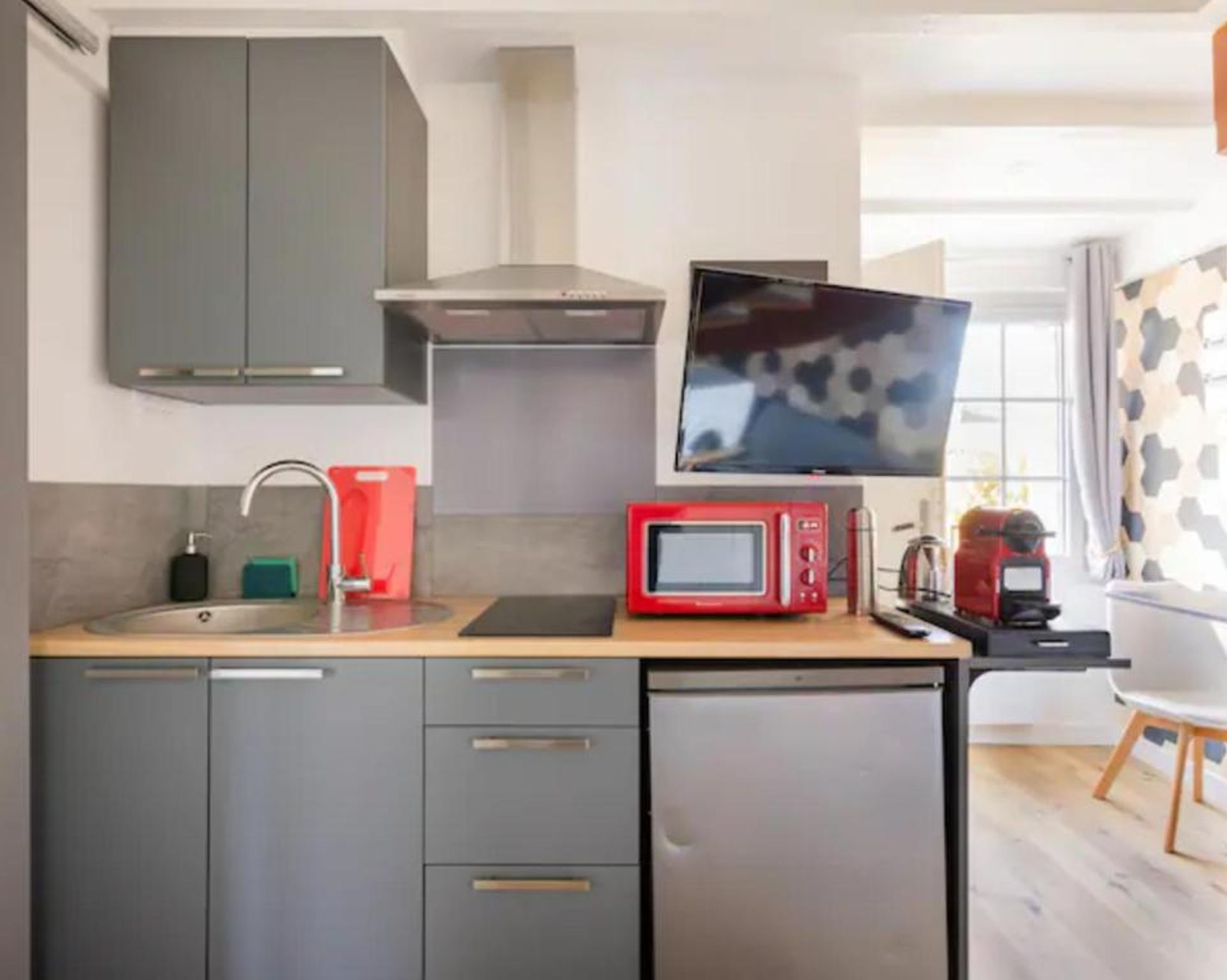 A compact kitchen with grey cabinets, a built-in microwave, a small refrigerator, a sink with a faucet, and a flat-screen TV mounted above the counter.