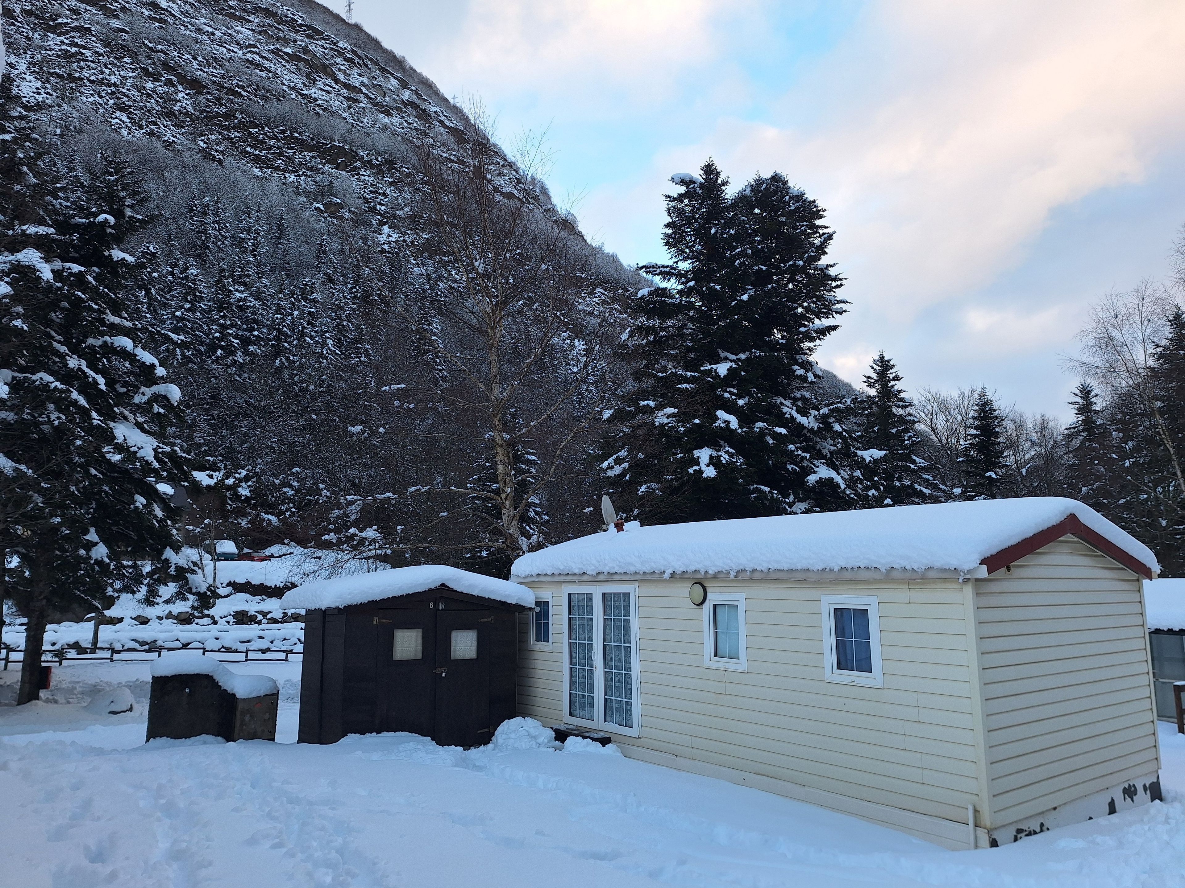 Une petite cabane à un seul étage, dont le toit est recouvert de neige, entourée d’arbres chargés de neige et avec une montagne en arrière-plan.