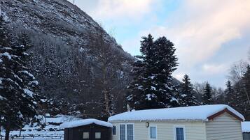 Une petite cabane à un seul étage, dont le toit est recouvert de neige, entourée d’arbres chargés de neige et avec une montagne en arrière-plan.