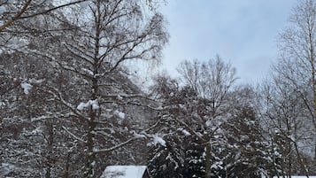 Lumipeitteinen maisema, jossa on puro, puita ja kivirakennus.