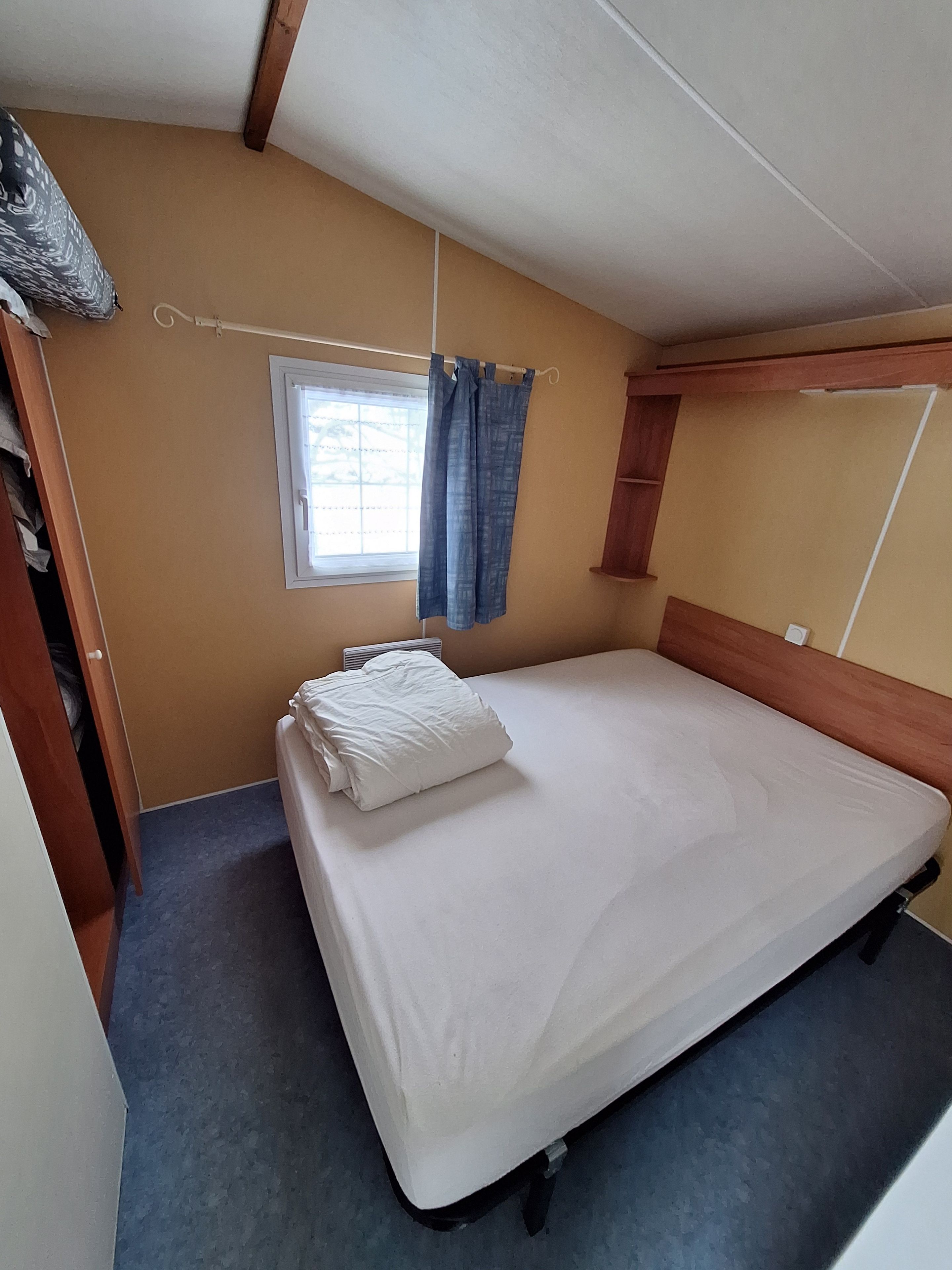 A small, single-bed room with a wooden headboard, a window with curtains, a radiator, and a small shelf.