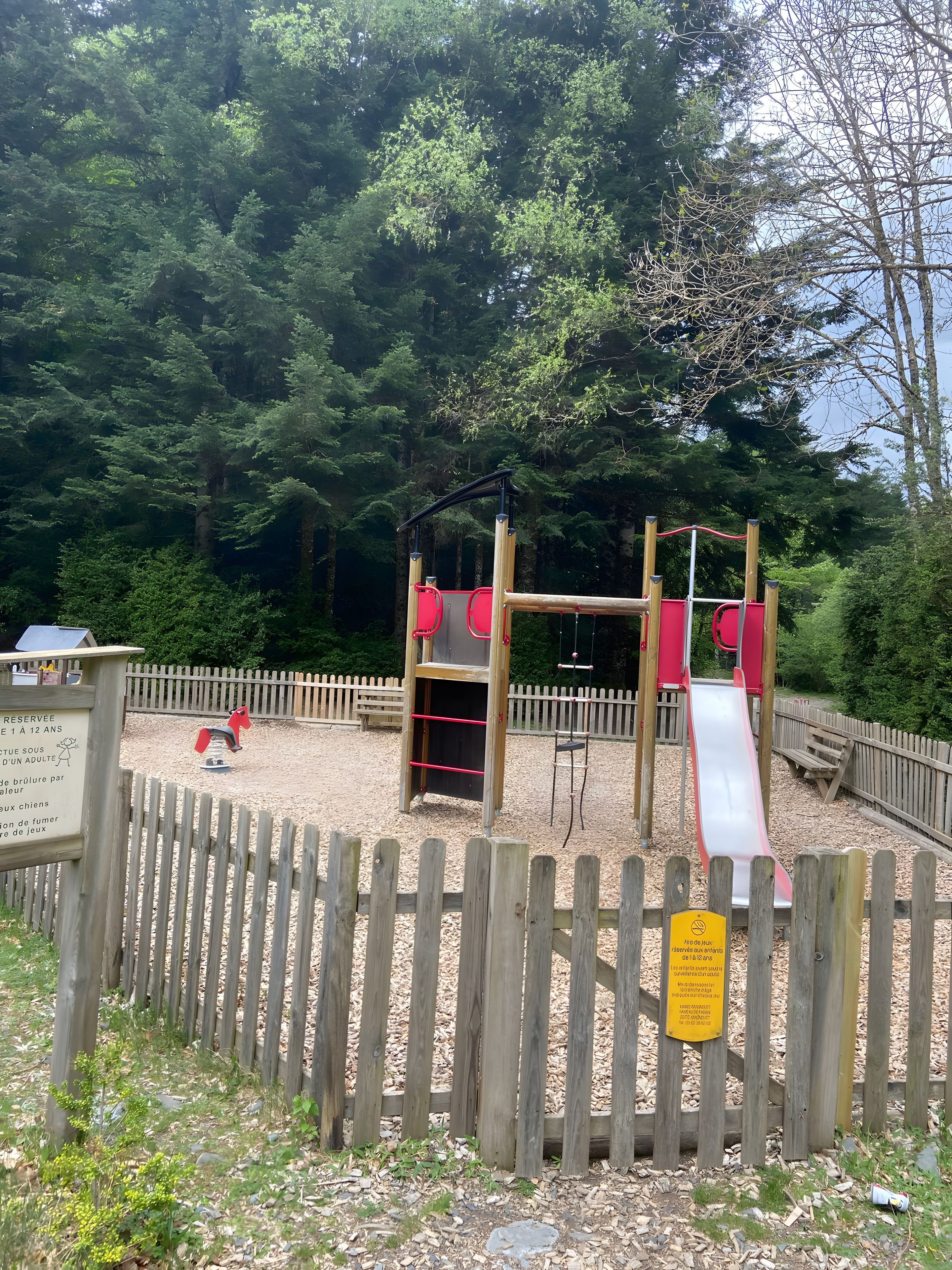 Une aire de jeux avec un toboggan et des structures d’escalade, entourée d’une clôture en bois et de nombreux arbres.
