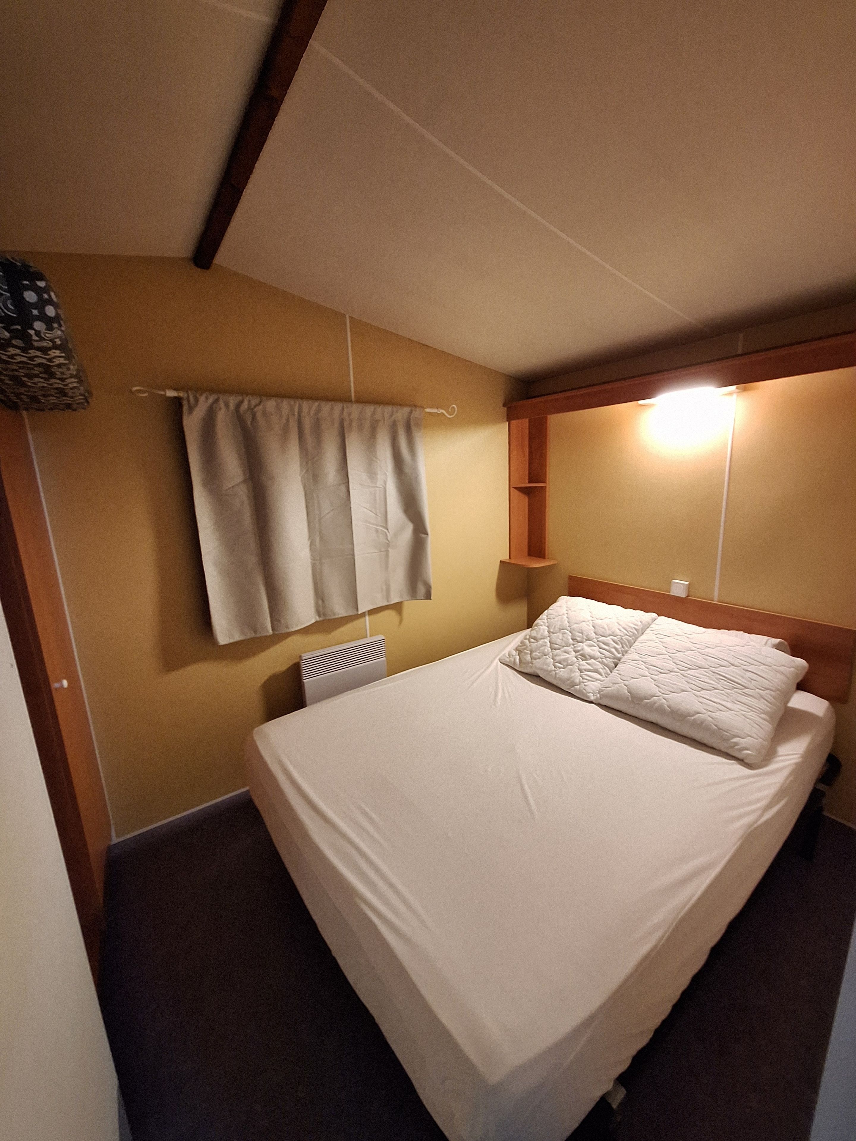 Un lit simple avec un matelas et des oreillers blancs, une table de chevet, une étagère fixée au mur, un radiateur et un rideau.