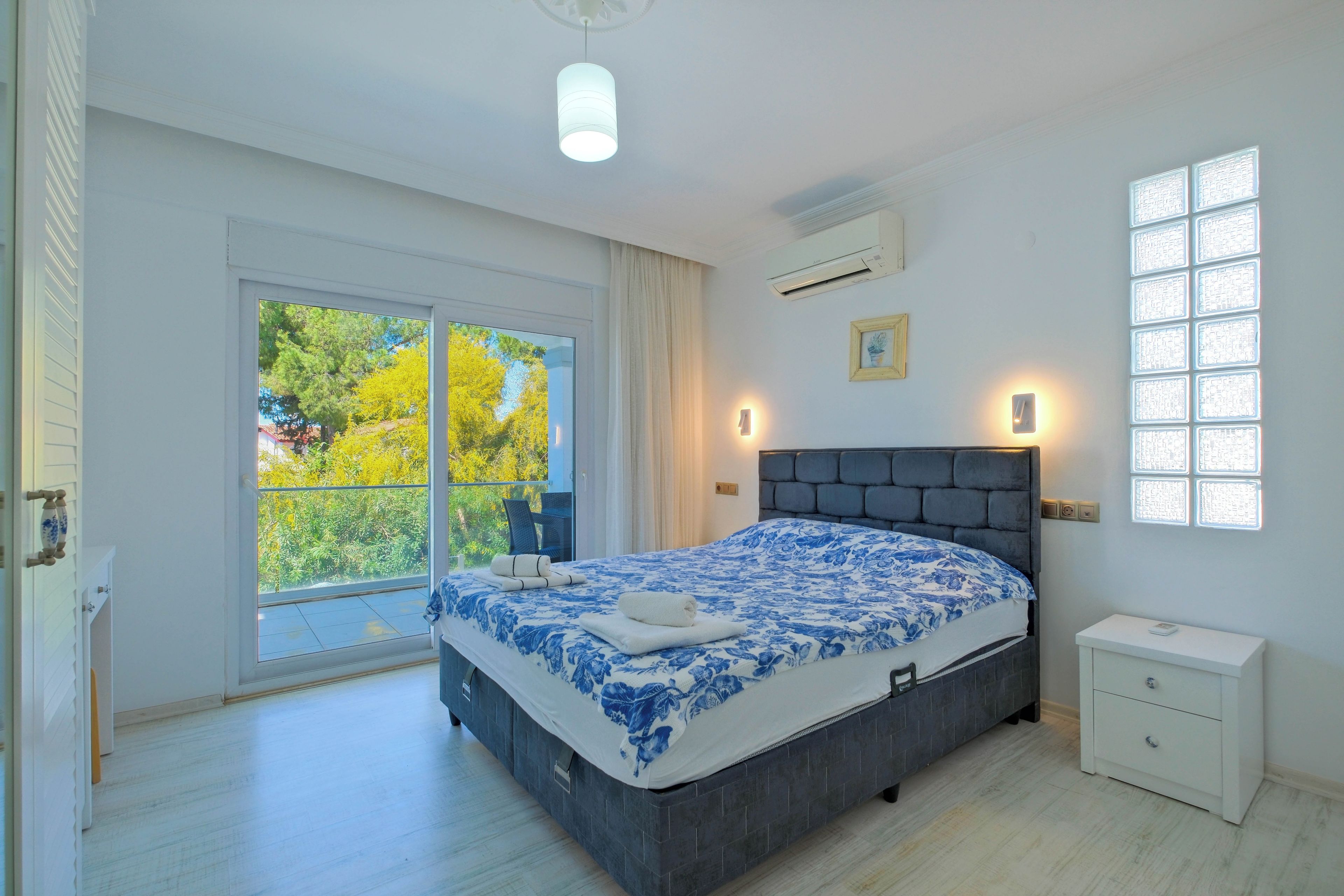A bedroom with a large bed, a window with a view of trees, a ceiling light, an air conditioner, and a bedside table.