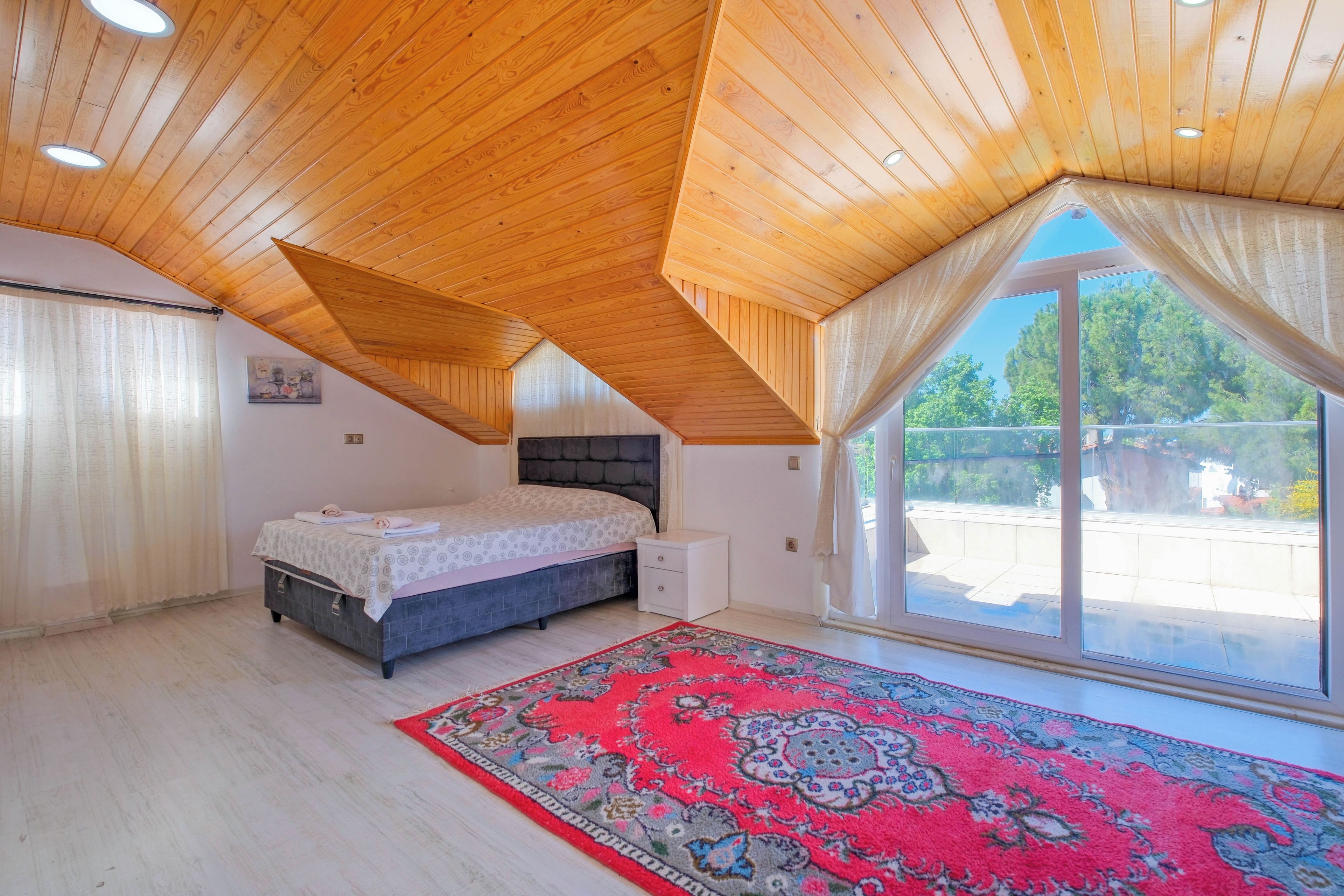 A bedroom with a bed, a nightstand, a window with curtains, and a red patterned rug.