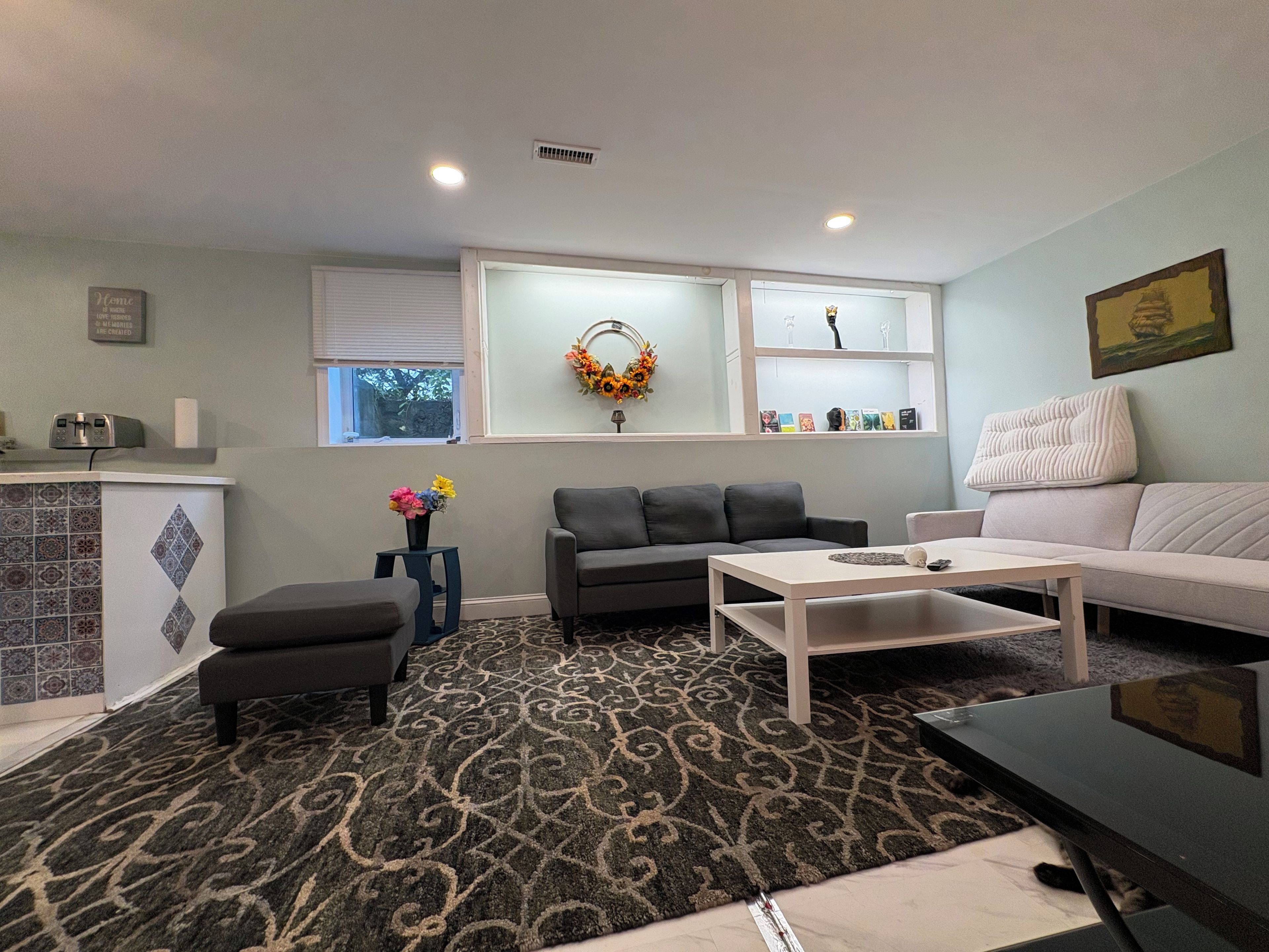 A living room with a grey sofa, a white coffee table, a patterned rug, a small side table with flowers, and a wall-mounted shelf with decorative items.