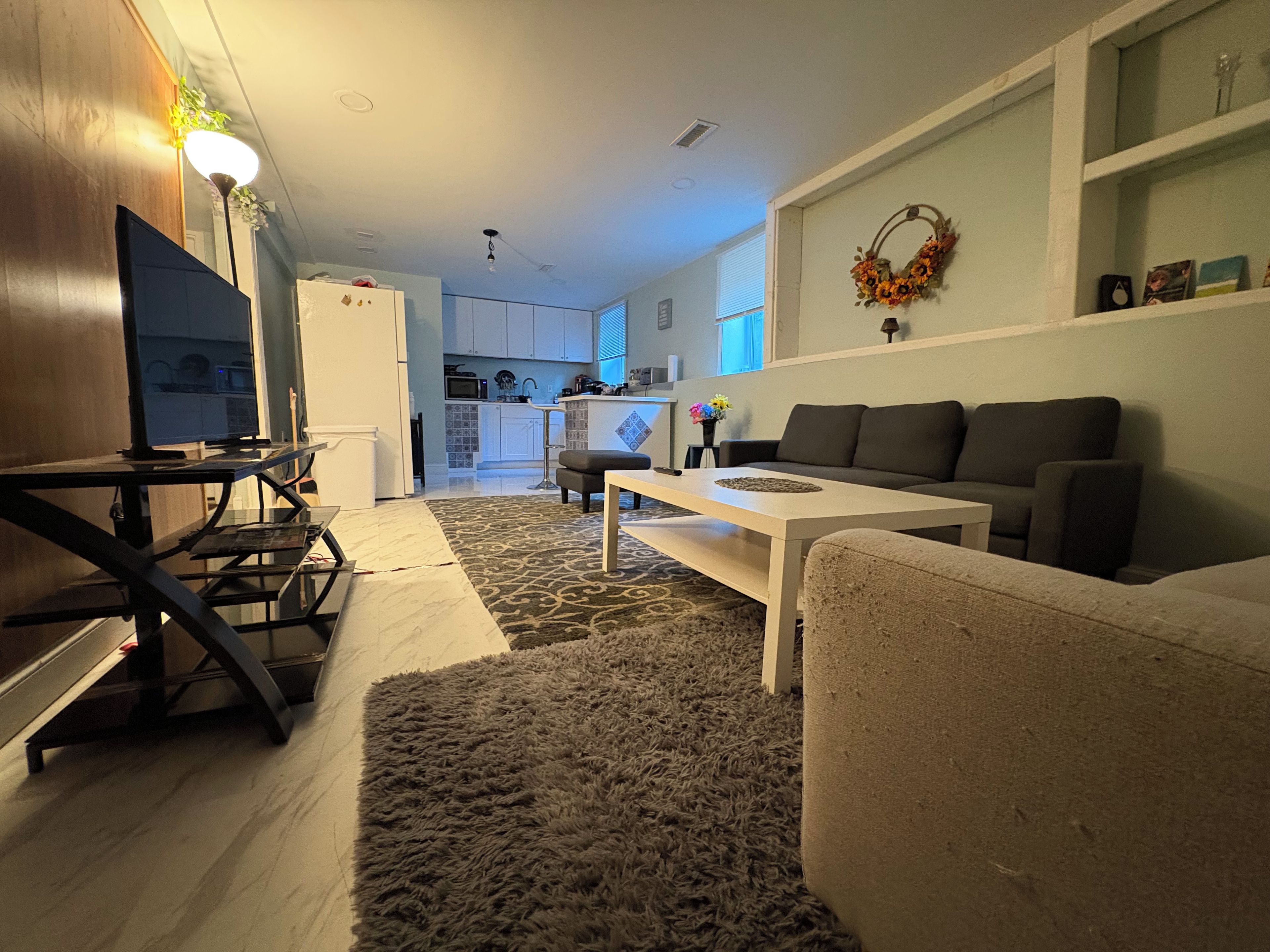 A modern living room with a sofa, coffee table, and TV. A kitchen is visible in the background.