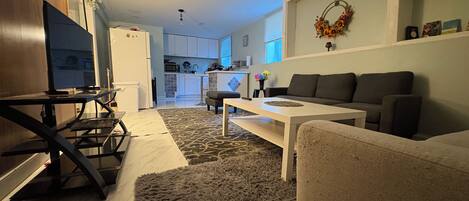 A modern living room with a sofa, coffee table, and TV. A kitchen is visible in the background.