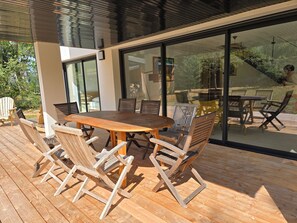 A wooden deck with a round table and chairs, large glass doors leading to an outdoor area with chairs and a table. - New Contemporary Home • Nature & Serenity • Direct Access to the GR34 (Crozon)