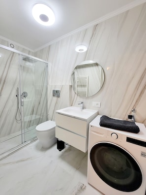 Ein modernes Badezimmer mit einer Glaskabine zur Dusche, einem weißen Waschbecken, einem Spiegel und einer Waschmaschine.