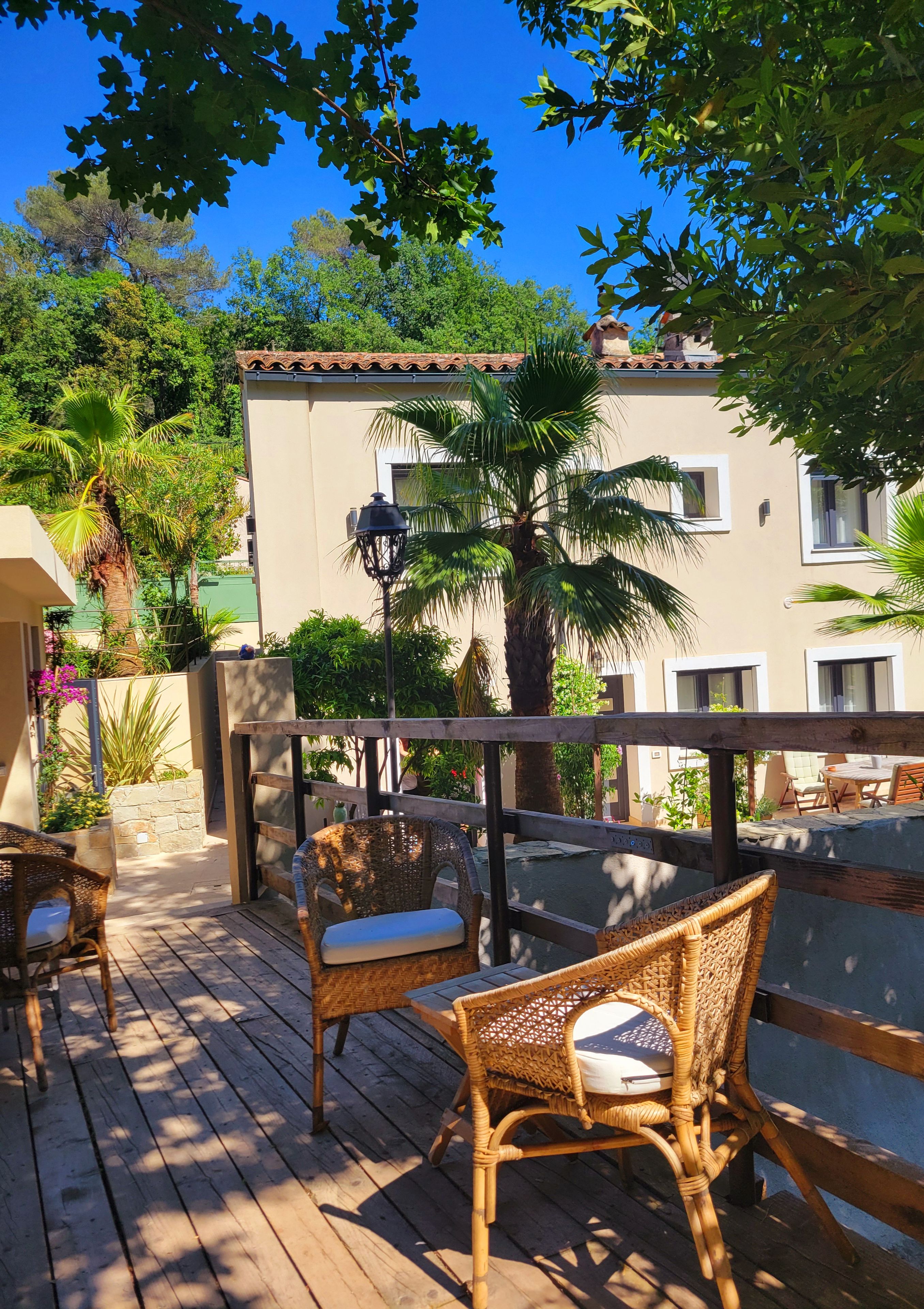 Une terrasse en bois avec des meubles en osier, un palmier et un bâtiment doté d’un toit en tuiles.
