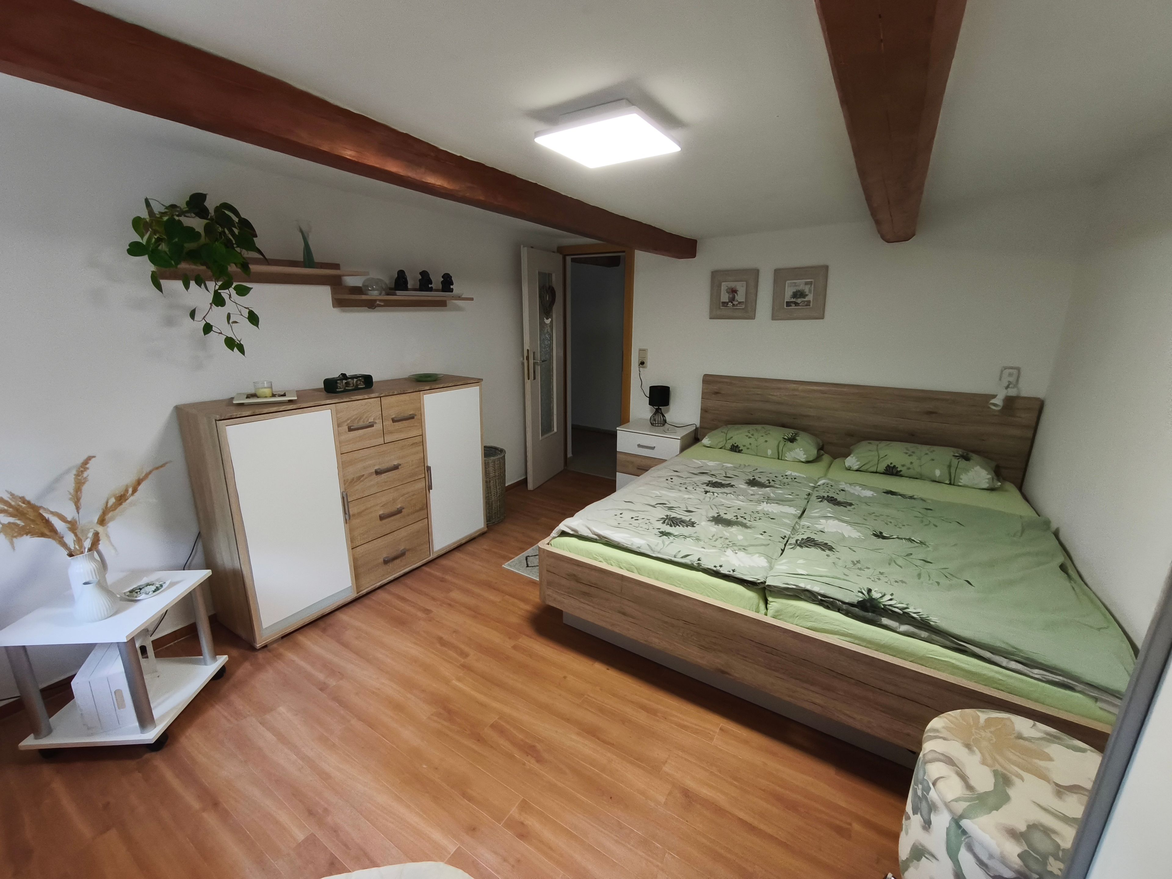 A bedroom with a wooden bed, a desk, a wardrobe, a nightstand, and a wall-mounted shelf with decorative items.