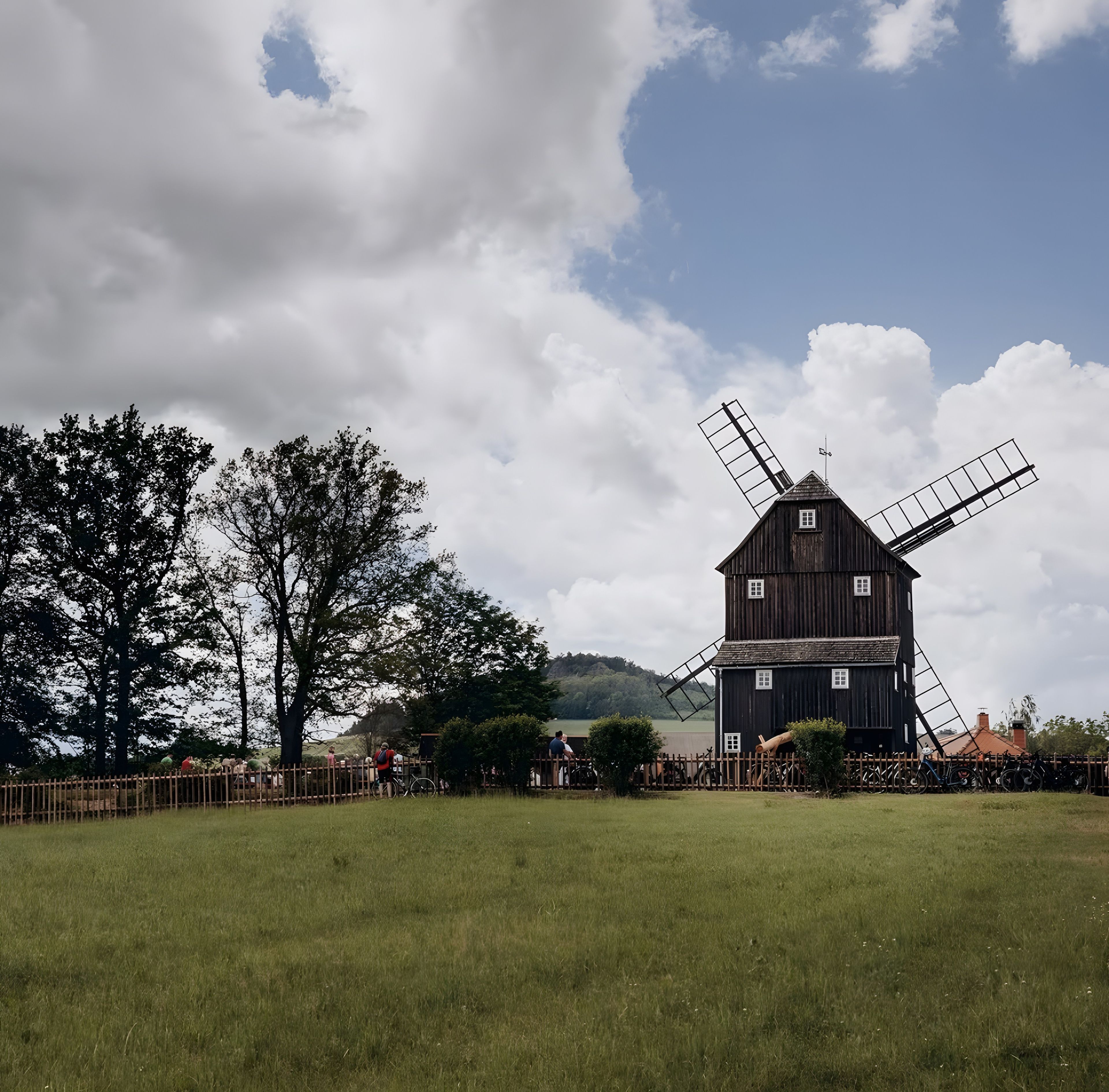 Un moulin à vent doté d’une structure en bois et d’une grande voile, entouré d’un champ herbeux et d’arbres.