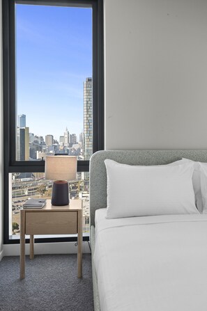 Quarto de hotel com cama, mesa de cabeceira e abajur. É visível uma paisagem urbana pela janela.