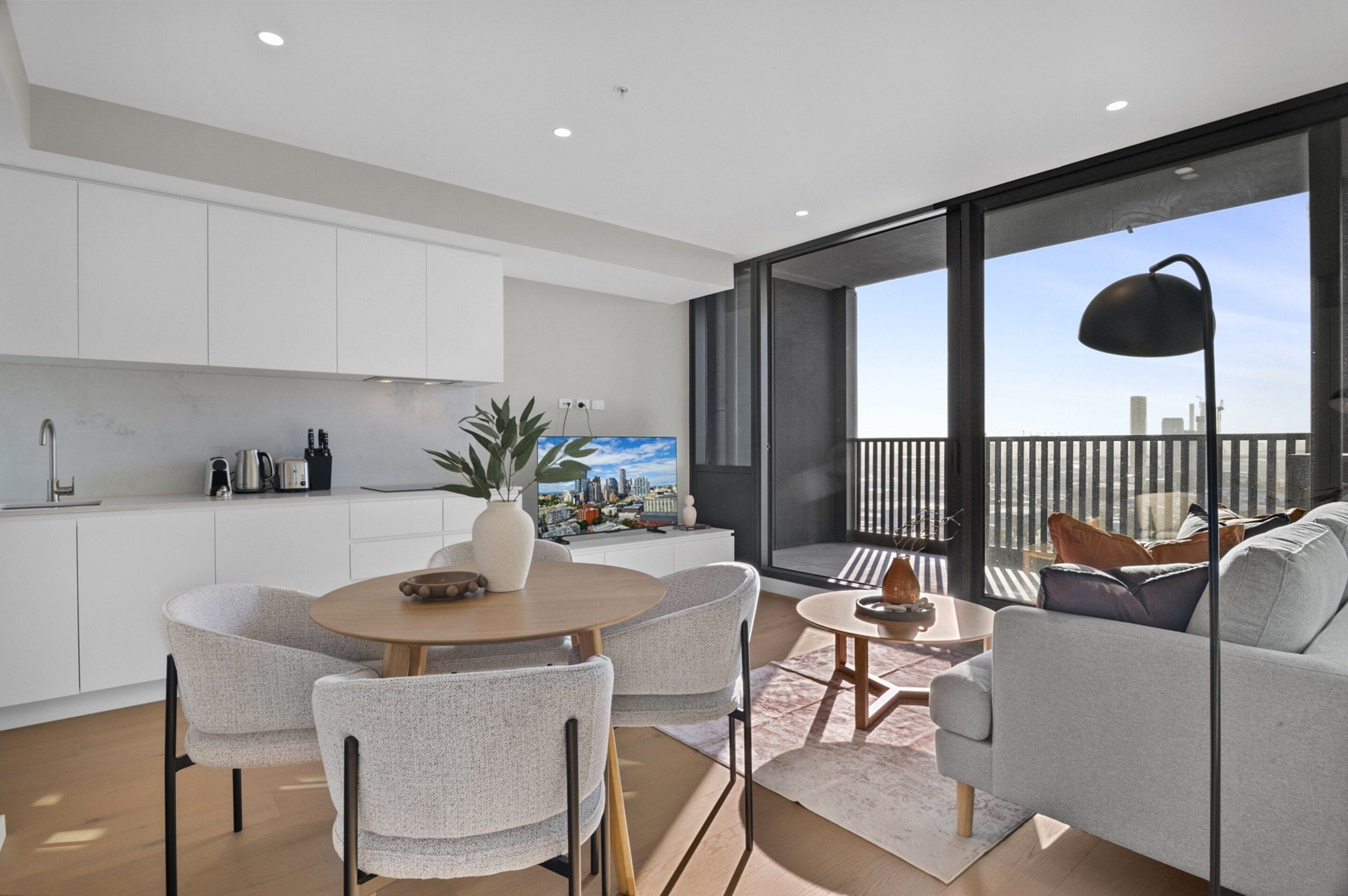 A modern kitchen with white cabinets, a dining area with a round table and chairs, and a balcony with a sofa and a city view.