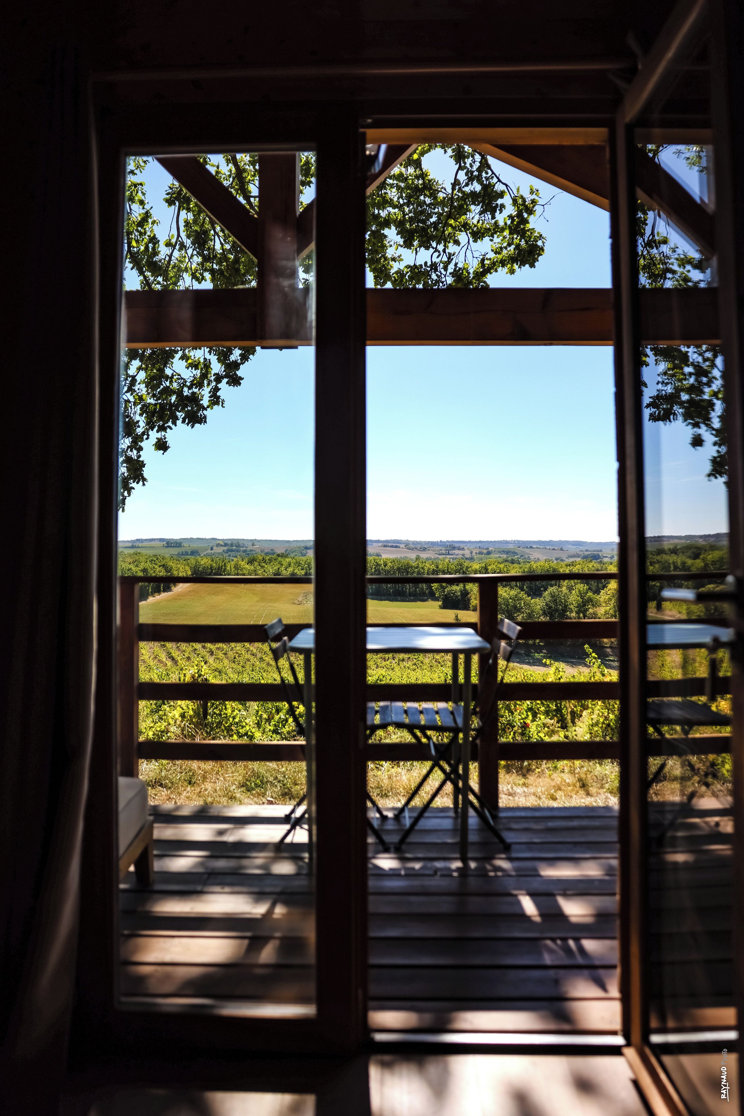 An open wooden door leading to an outdoor deck with a table and chairs, overlooking a green landscape.
