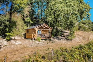Eine Holzhütte mit Veranda, umgeben von Bäumen und einer trockenen, grasbewachsenen Fläche.