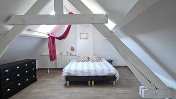 Ein Schlafzimmer mit schrägen Wänden, einem Bett mit gemustertem Überwurf, einem Schminktisch und einem Fenster mit Vorhang.