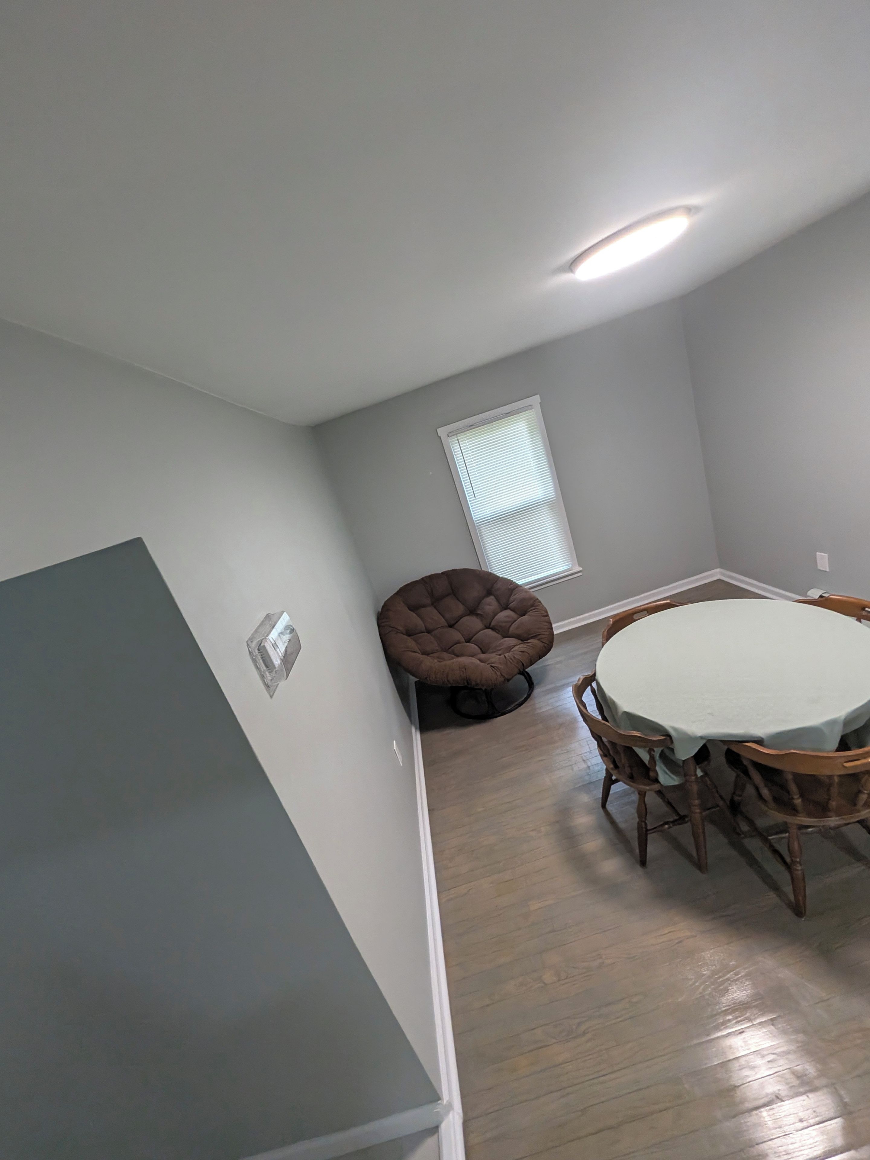 A dining area with a round table and chairs, a tufted chair, and a window with blinds.