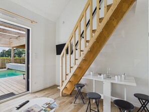 Un interior moderno con una escalera de madera, una piscina visible a través de un ventanal y una mesa blanca con taburetes de bar.