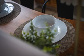 Uma mesa com uma xícara e pires brancos sobre um jogo americano, com uma planta verde desfocada em primeiro plano.