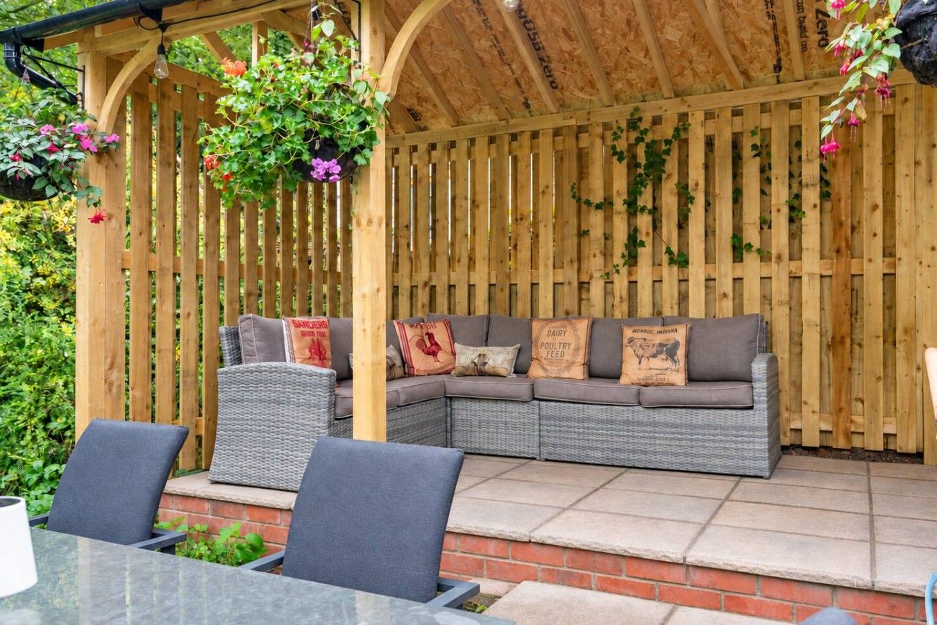 A covered outdoor seating area with a wooden pergola, a grey sofa with decorative pillows, and hanging plants.