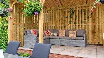 A covered outdoor seating area with a wooden pergola, a grey sofa with decorative pillows, and hanging plants.