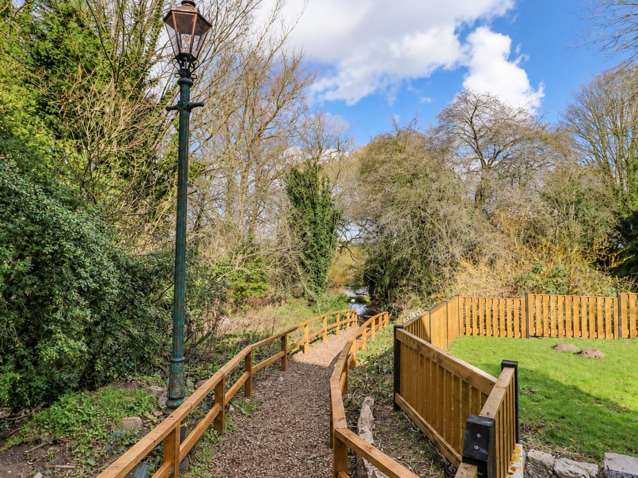 A wooden pathway leading to a bridge over a stream, surrounded by trees and a wooden fence.