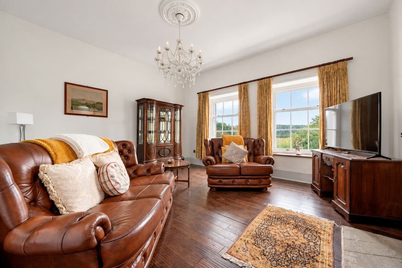 A spacious living room with a leather sofa, a wooden cabinet, a television, and a large window with curtains.