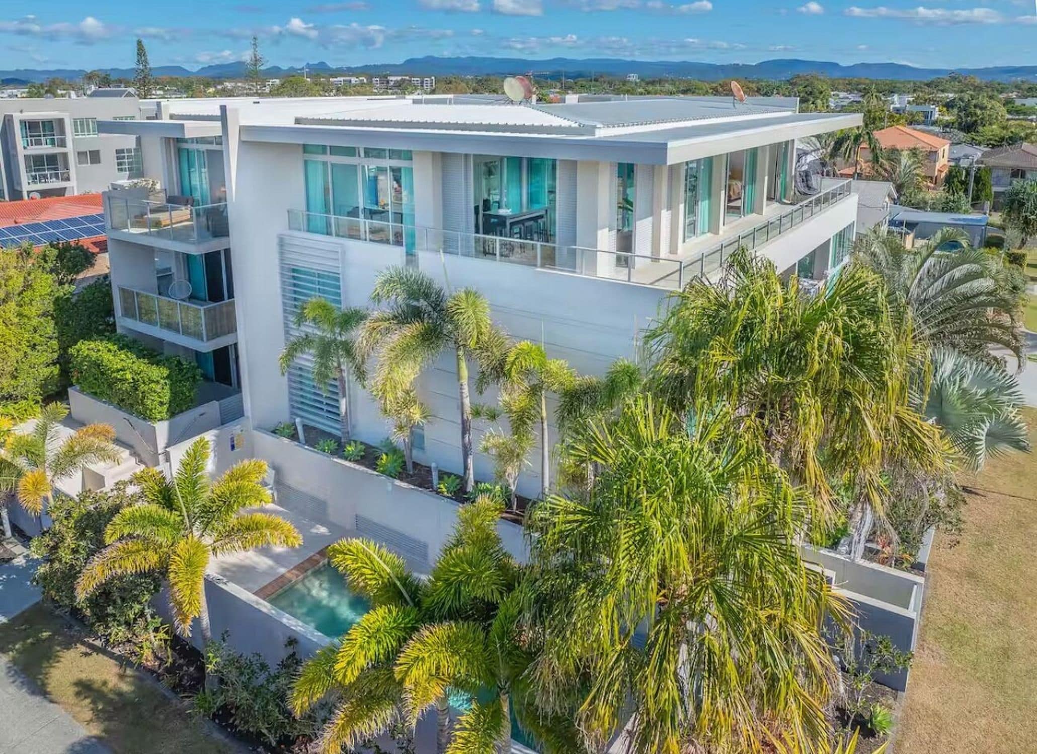 A modern multi-story residential building with a pool, surrounded by palm trees and a clear view of the landscape.