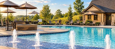 Una piscina con múltiples chorros de agua, rodeada de sillas de descanso y sombrillas, con un edificio y árboles al fondo.