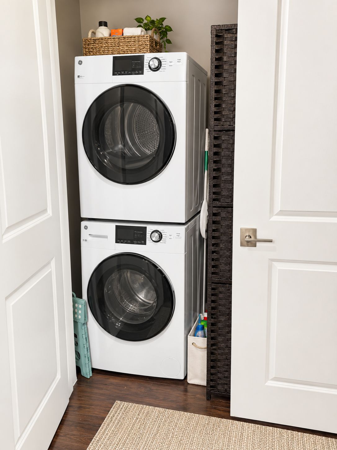A laundry room with two stacked front-loading washing machines, a wicker basket on top, and a door with a dark patterned frame.