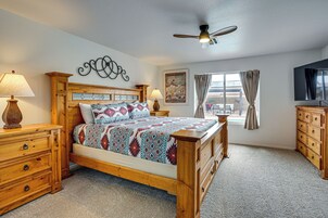 A bedroom with a wooden bed, two bedside tables with lamps, a ceiling fan, a window with curtains, and a wall-mounted TV.
